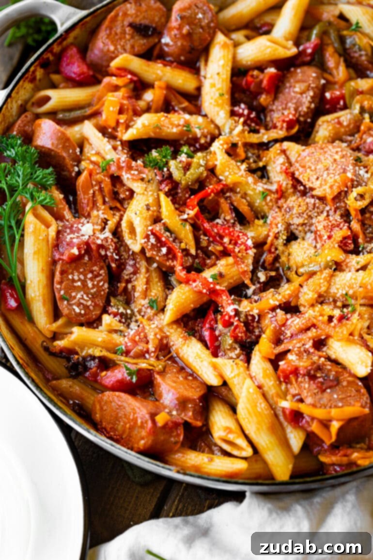 a photo of cooked penne pasta in a skillet mixed with sauteed peppers and onions and sausage.