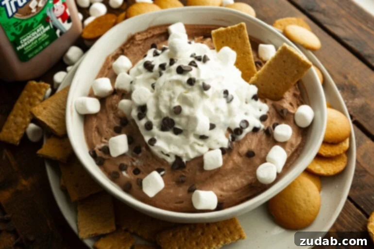 Festive Holiday Hot Chocolate Dip 3 A large white bowl of hot cocoa dip topped with whipped cream and sprinkled with mini chocolate chips and mini marshmallows, with two graham crackers sticking out of it.