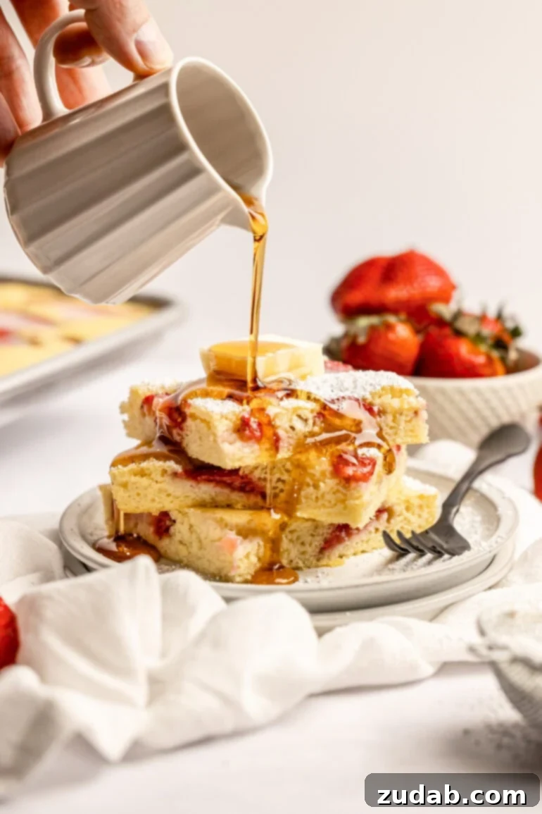 Sheet Pan Strawberry Pancake Bake 13 A hand gently pouring rich maple syrup over a freshly cut stack of strawberry sheet pan pancakes, ready to be savored for breakfast or brunch.