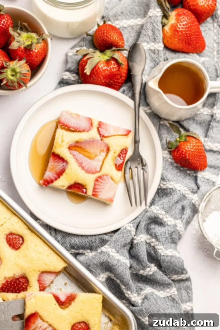 Sheet Pan Strawberry Pancake Bake 14 A serving of strawberry sheet pan pancakes on a small plate, generously topped with maple syrup. A full sheet pan of the baked pancakes and a bottle of syrup are visible in the background, showcasing the abundance and ease of this breakfast solution.
