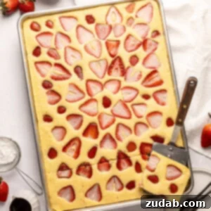 A half sheet pan full of baked sheet pan pancakes topped with slices of fresh strawberry baked into the pancakes and a metal spatula removing one serving