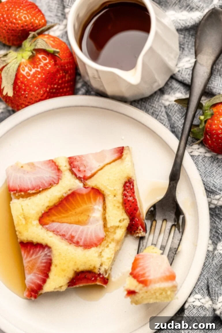 Sheet Pan Strawberry Pancake Bake 3 A single slice of fluffy strawberry sheet pan pancake rests on a white plate, with a corner delicately removed by a fork, hinting at its tender texture. A bottle of maple syrup sits nearby, ready for drizzling.
