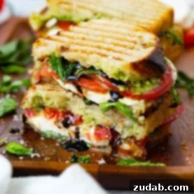 A grilled caprese panini cut in half, showing arugula, tomatoes, pesto, and mozzarella cheese inside, with balsamic glaze oozing out.