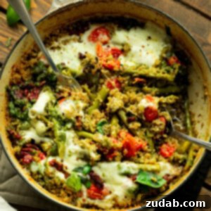 A cast iron skillet of quinoa mixed with pesto, asparagus, tomatoes, and topped with melted cheese.