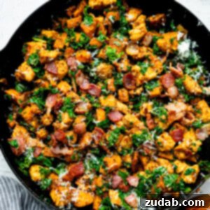 A cast iron skillet full of golden chunks of chicken mixed with bacon crumbles, kale, and sweet potatoes.