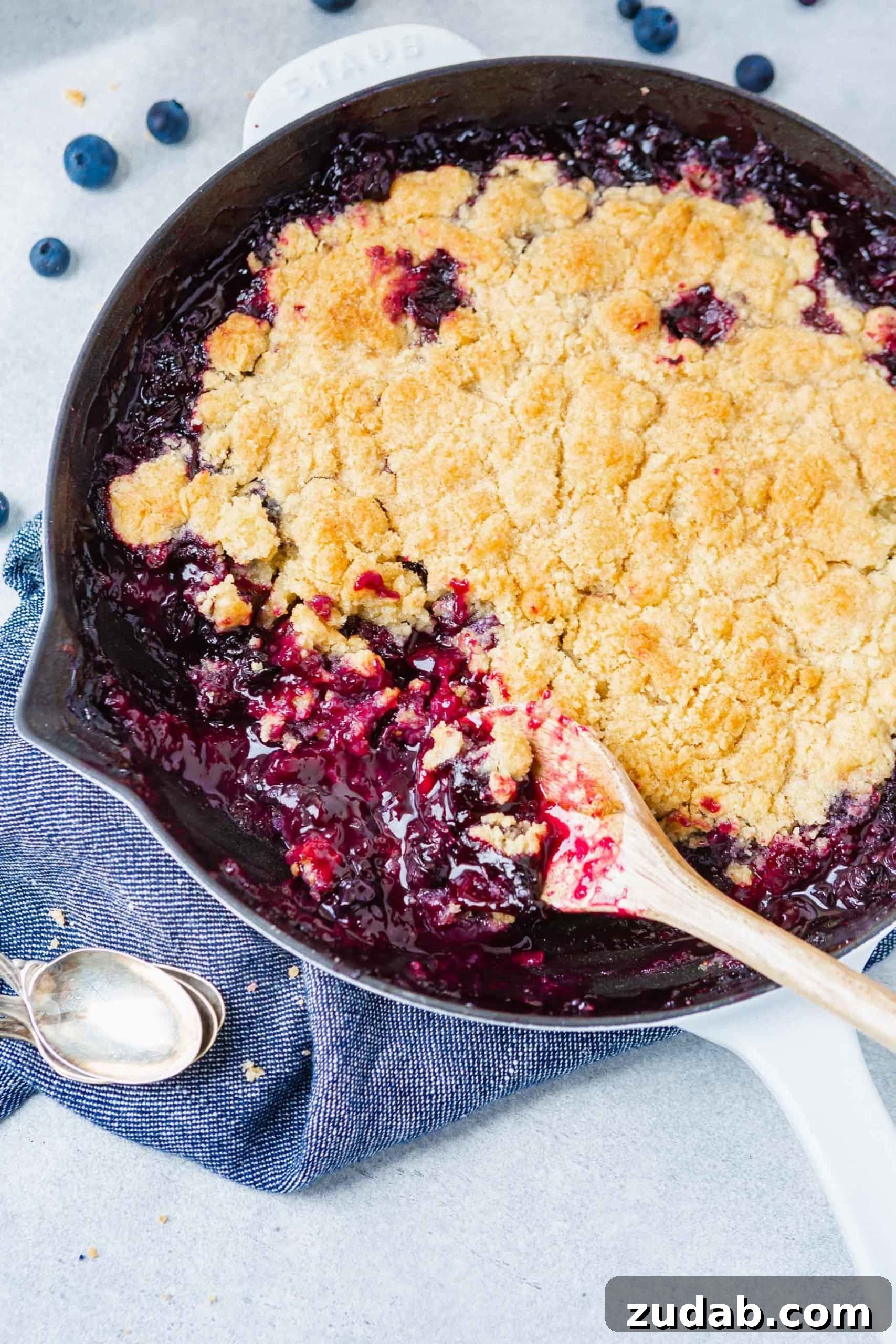 Summer Blueberry Cobbler A photo of a blueberry cobbler in a cast iron skillet with a golden topping and a serving being taken out with a wooden spoon.