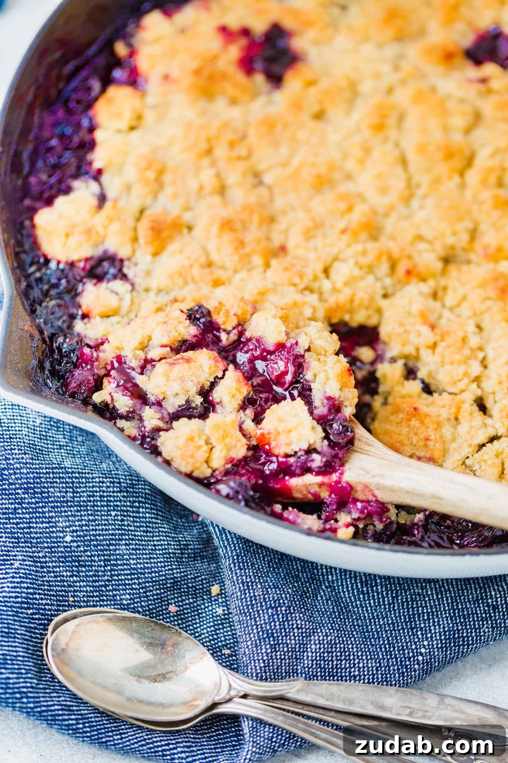 Summer Blueberry Cobbler A photo of a blueberry cobbler with golden topping in a cast iron skillet. A wooden spoon is scooping out a serving.