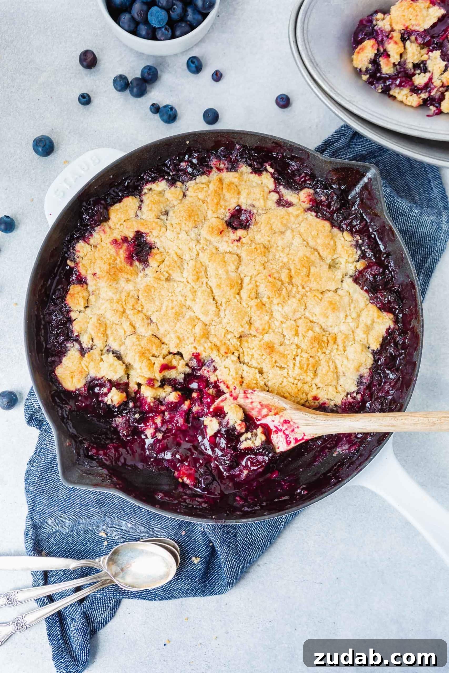 Summer Blueberry Cobbler A cast iron skillet full of blueberry cobbler with a golden topping. A wooden spoon is scooping out a serving.