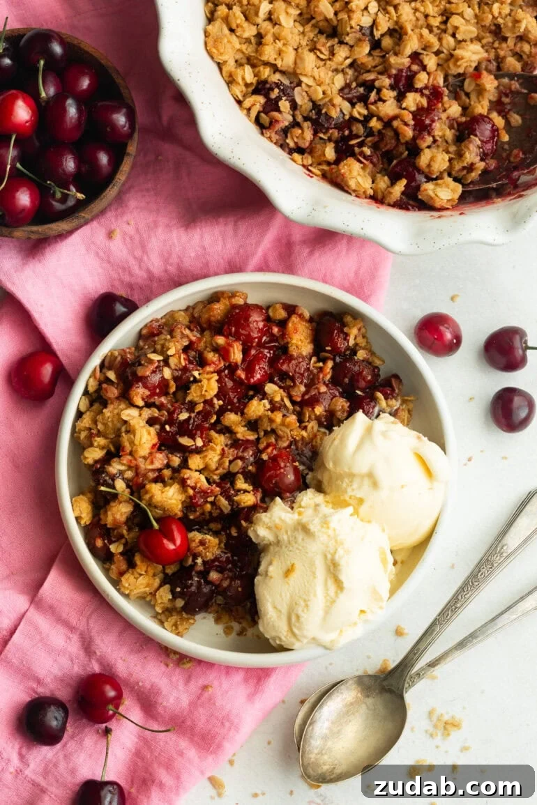 Classic Old-Fashioned Cherry Crisp with Oat Topping 4 a serving of cherry crisp on a small dessert plate with two scoops of vanilla on the side and the whole cherry crisp in a baking dish sitting next to the plate